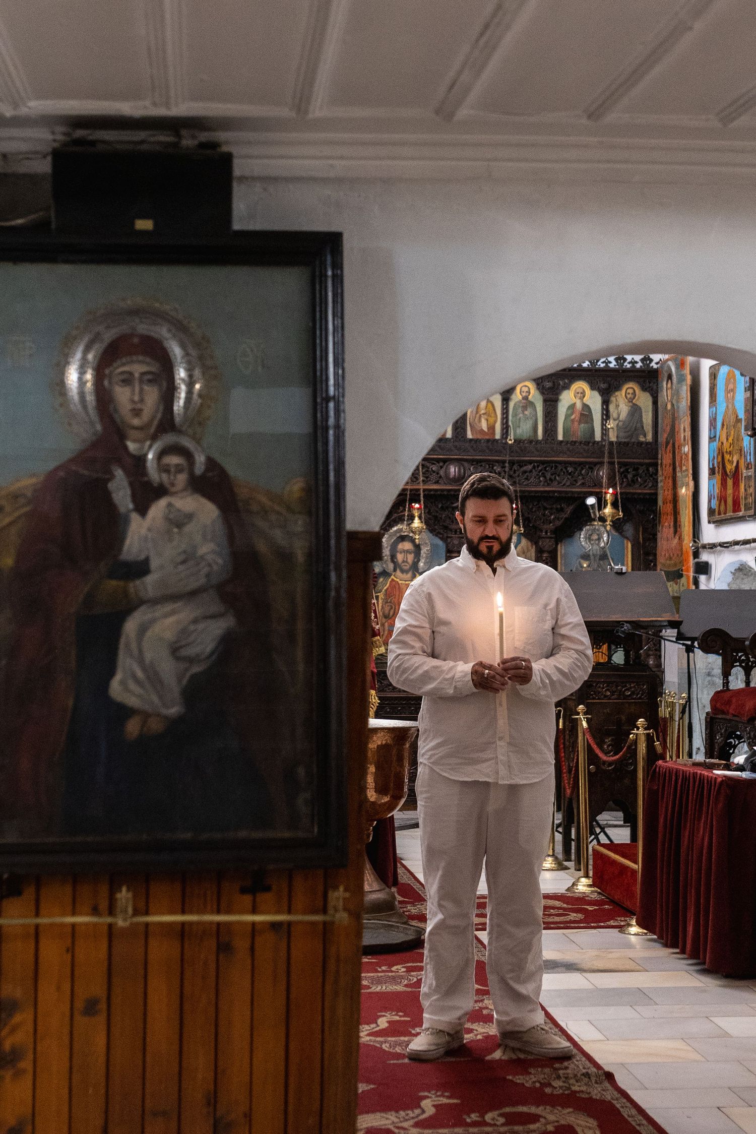 Man in white holding candle before Orthodox icon of Virgin Mary and Child in decorated church interior.