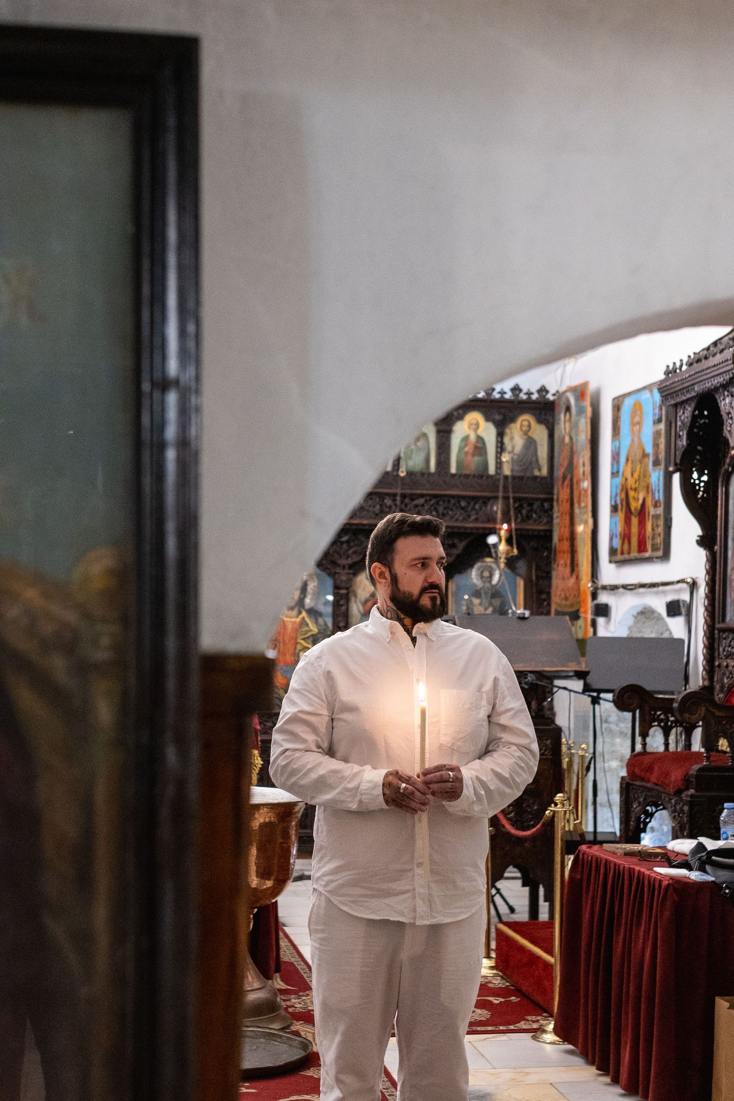 Man in white attire holding a lit candle inside an Orthodox church with icons and ornate decor.