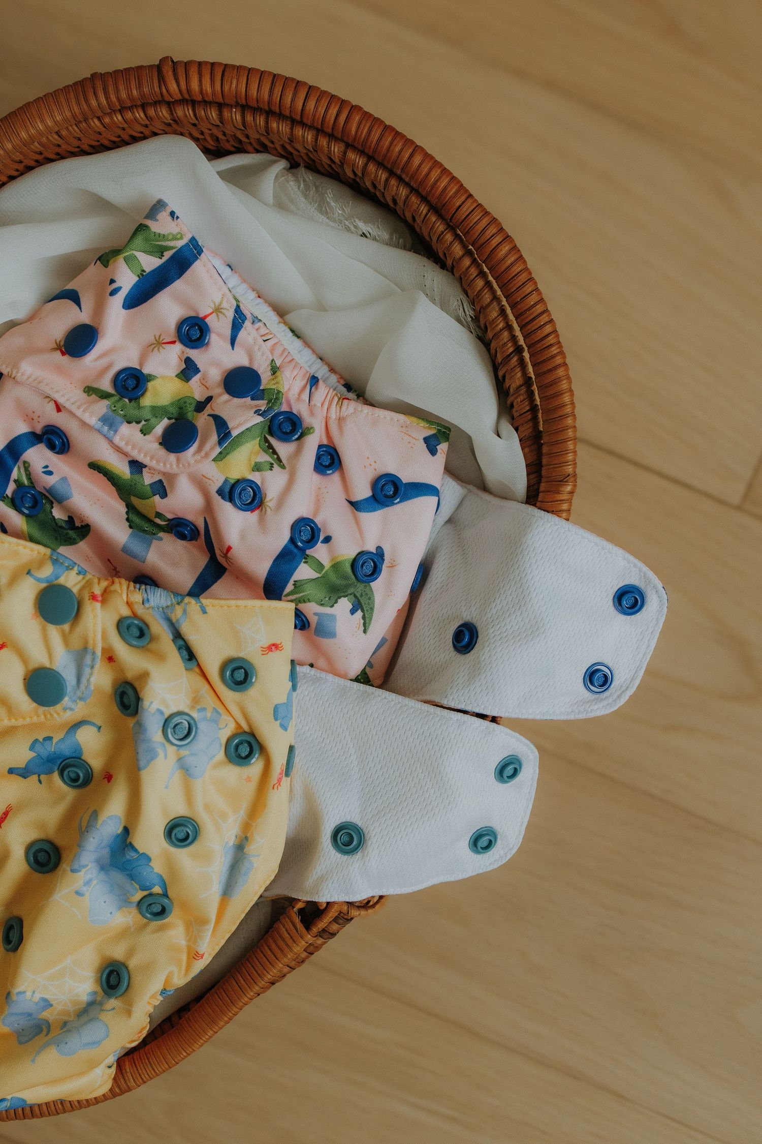 Colorful cloth diapers with snap closures in a wicker basket, featuring playful animal prints on wood floor.