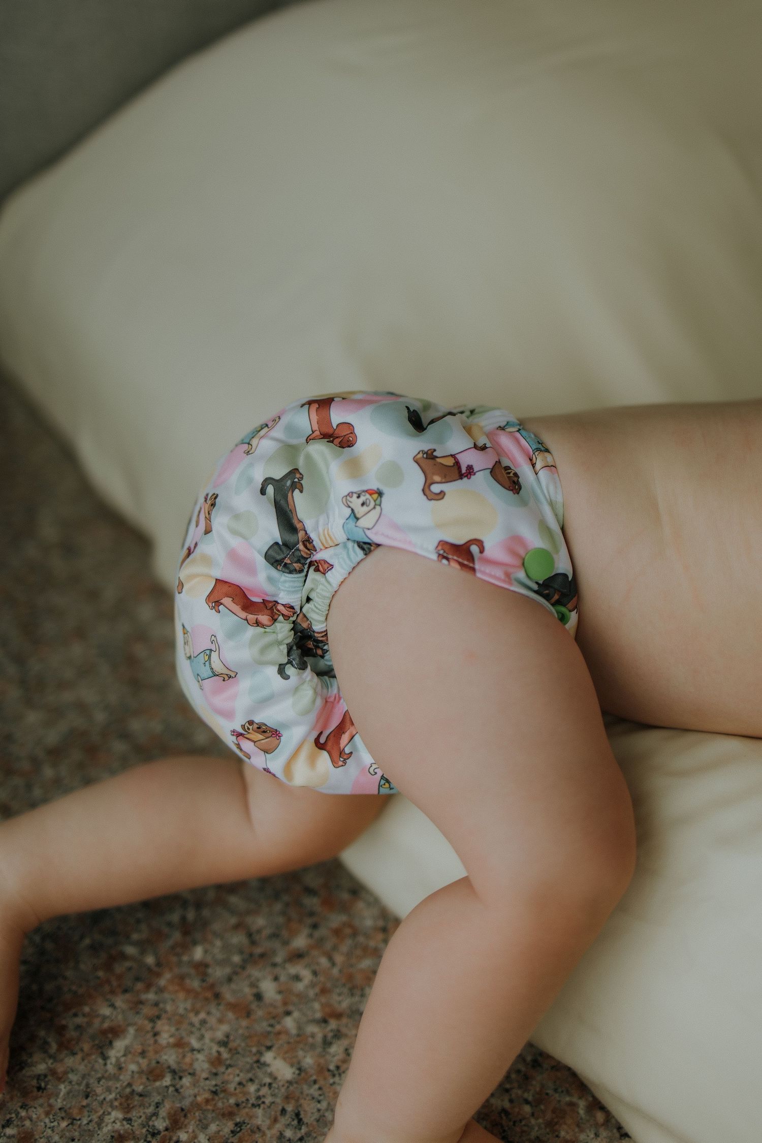 Baby wearing a colorful printed cloth diaper with playful animal patterns, crawling on a light-colored cushioned surface.