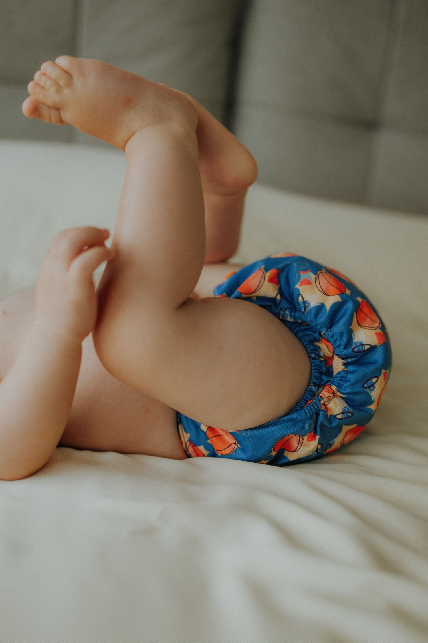 Baby wearing a colorful blue cloth diaper with bird print, lying on a white bed with legs raised.