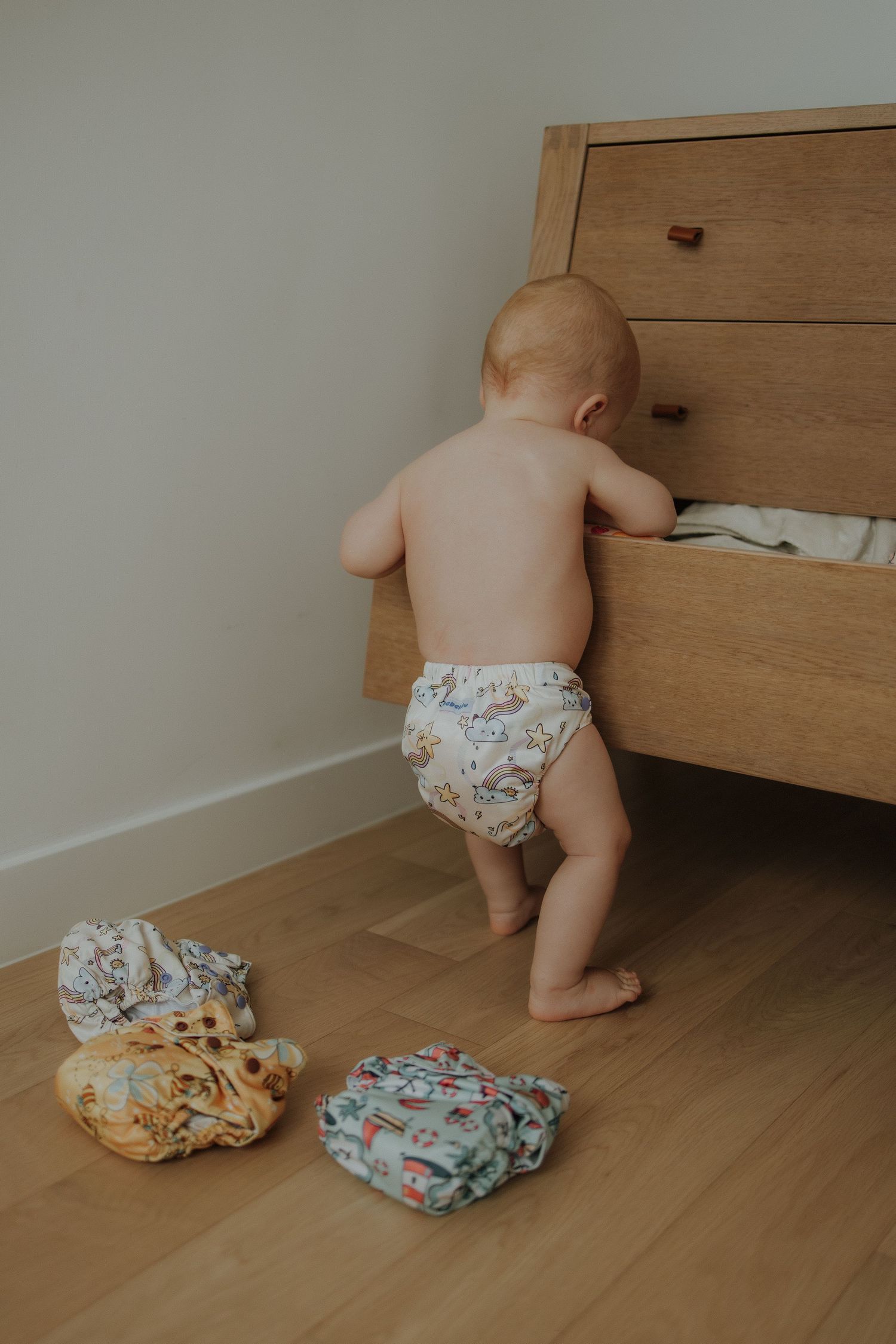 Baby in cloth diaper standing at wooden dresser drawer with colorful reusable diapers on floor.