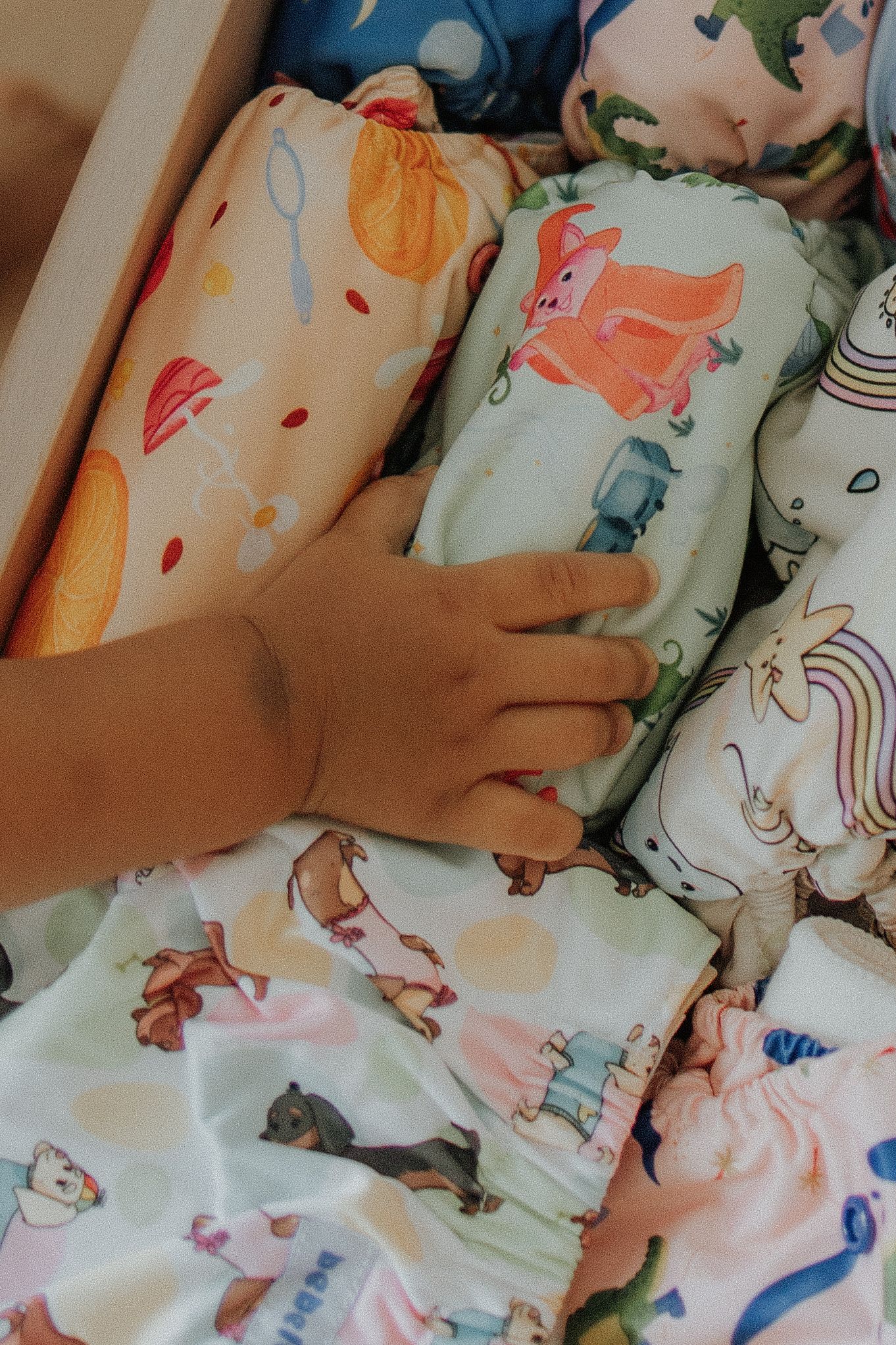 Baby hand reaching into a drawer of colorful cloth diapers with fun animal and unicorn prints.