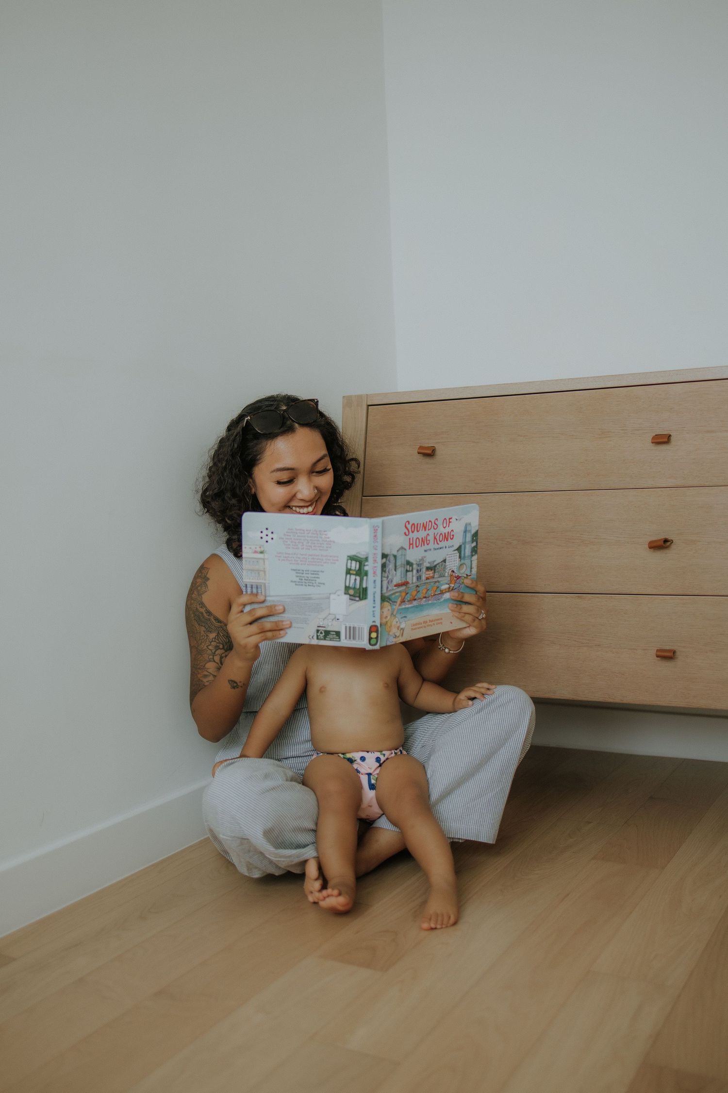 Smiling mother with tattoos sits on floor reading colorful picture book with toddler in patterned diaper cover.