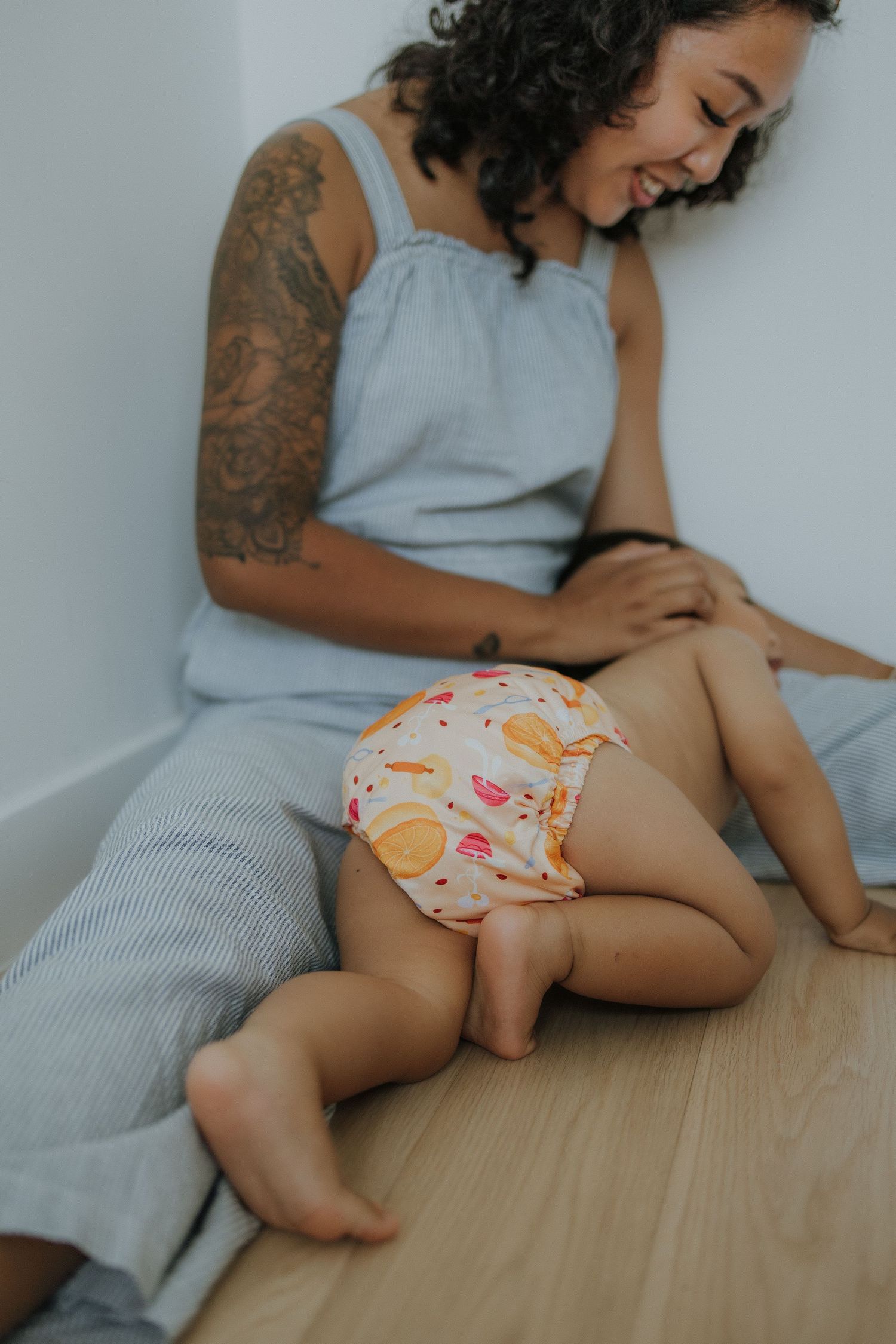 Smiling tattooed mother sitting on floor with baby in colorful diaper crawling nearby.