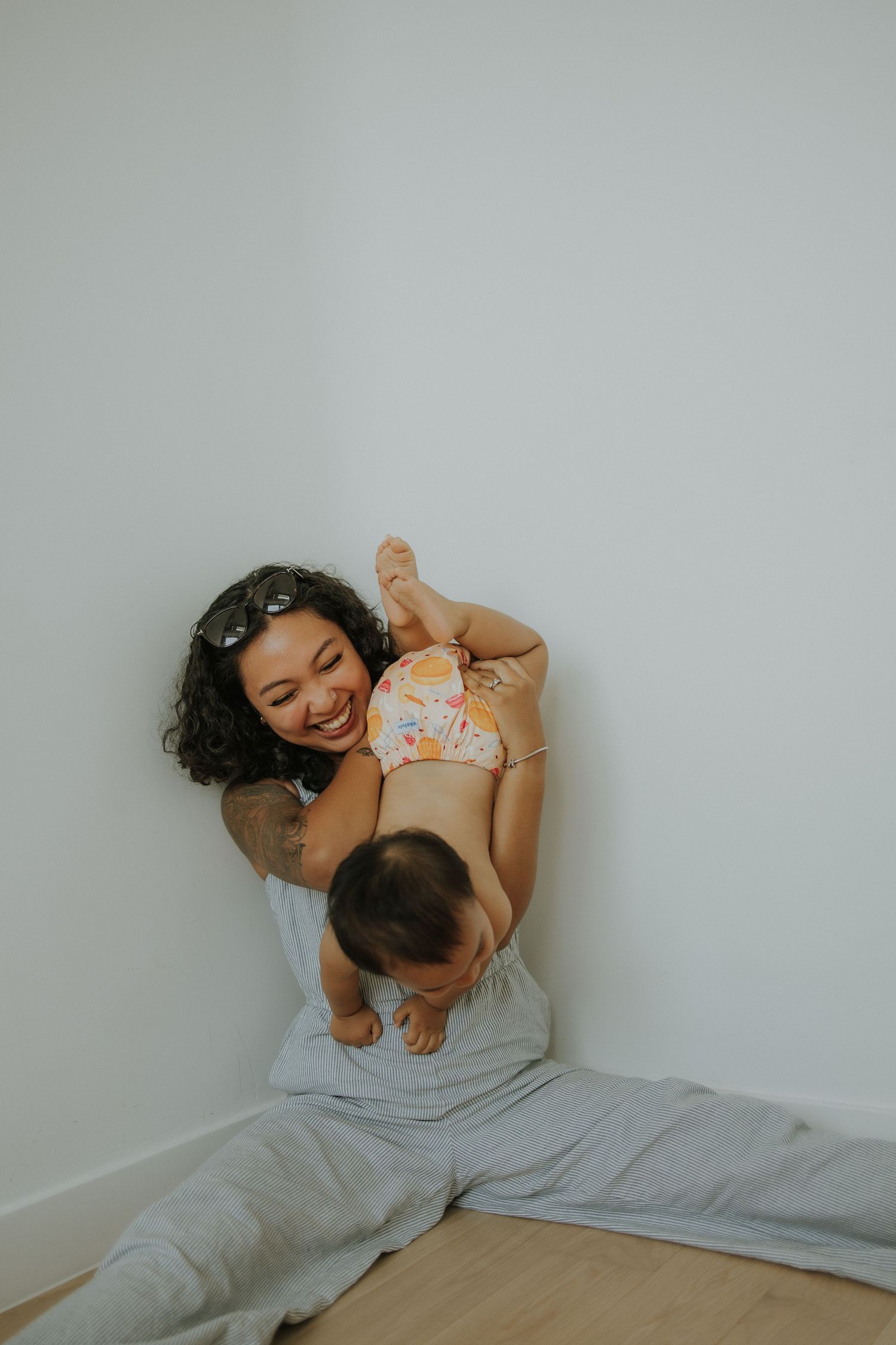 A smiling mother sits against a white wall, joyfully lifting her baby in a colorful outfit while seated on a gray blanket.