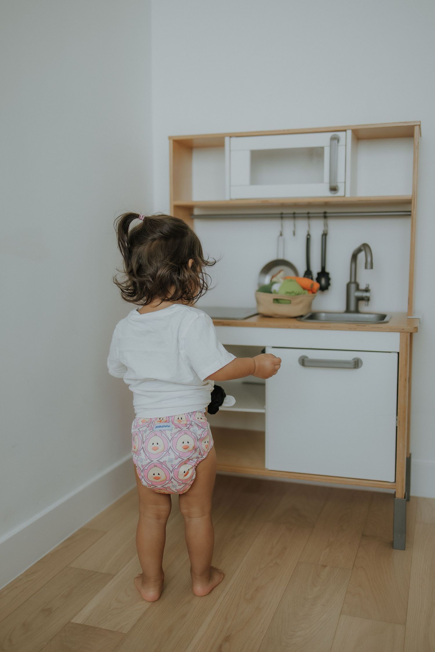 Toddler girl playing with IKEA DUKTIG wooden toy kitchen set, wearing white shirt and colorful diaper.
