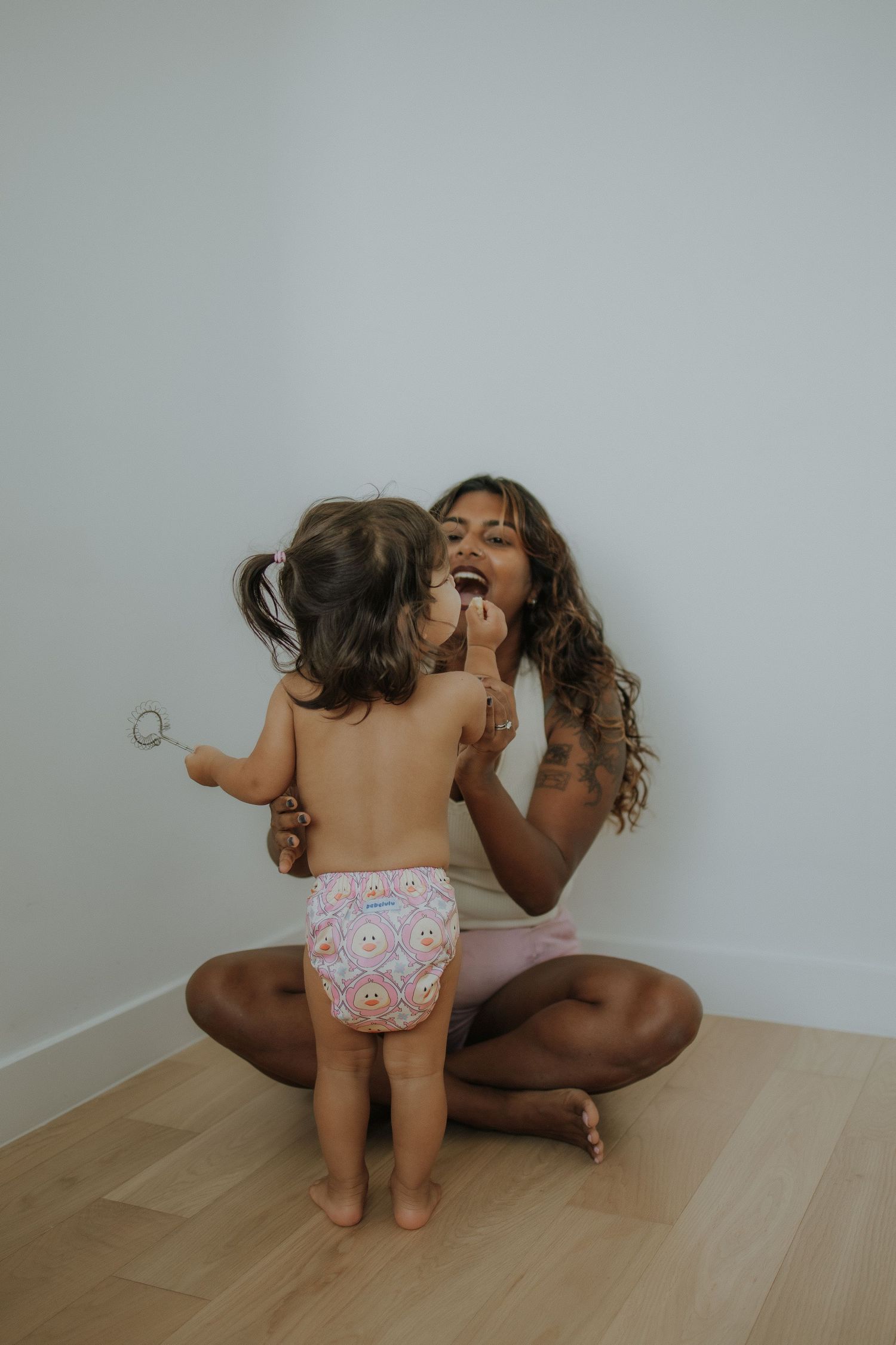 A mother sits cross-legged on a wood floor as her toddler in a floral diaper applies makeup to her face.