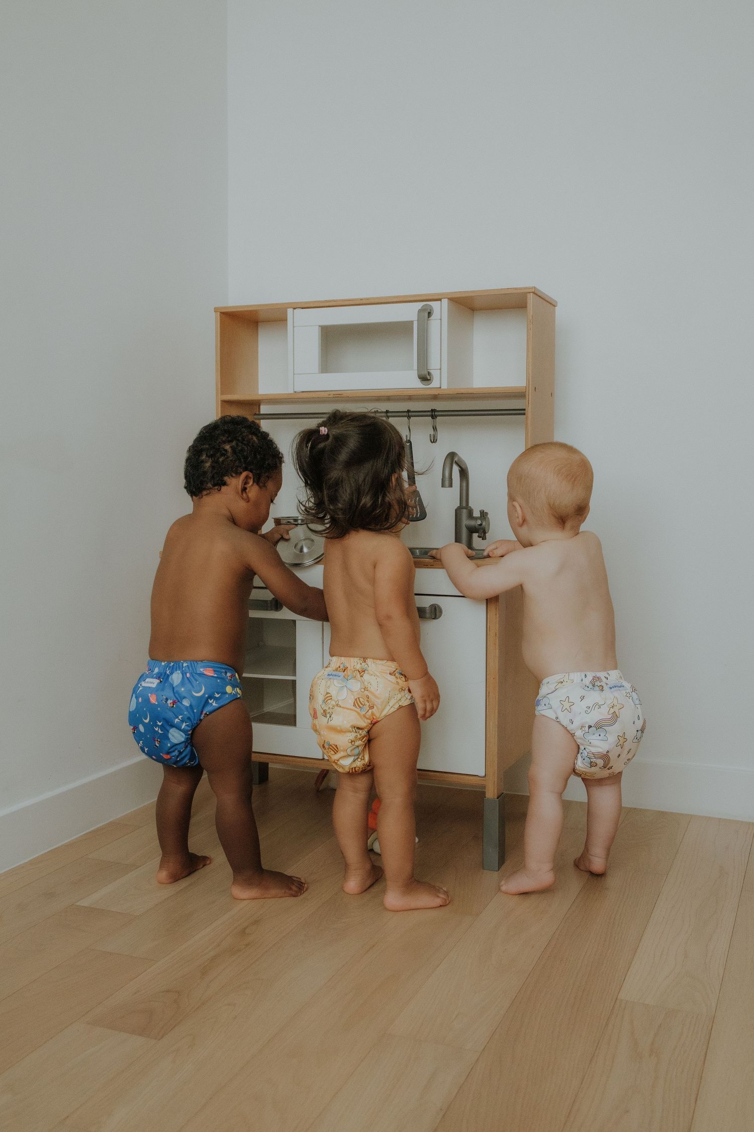 Three toddlers in colorful cloth diapers playing together at a wooden toy kitchen set on hardwood floors.