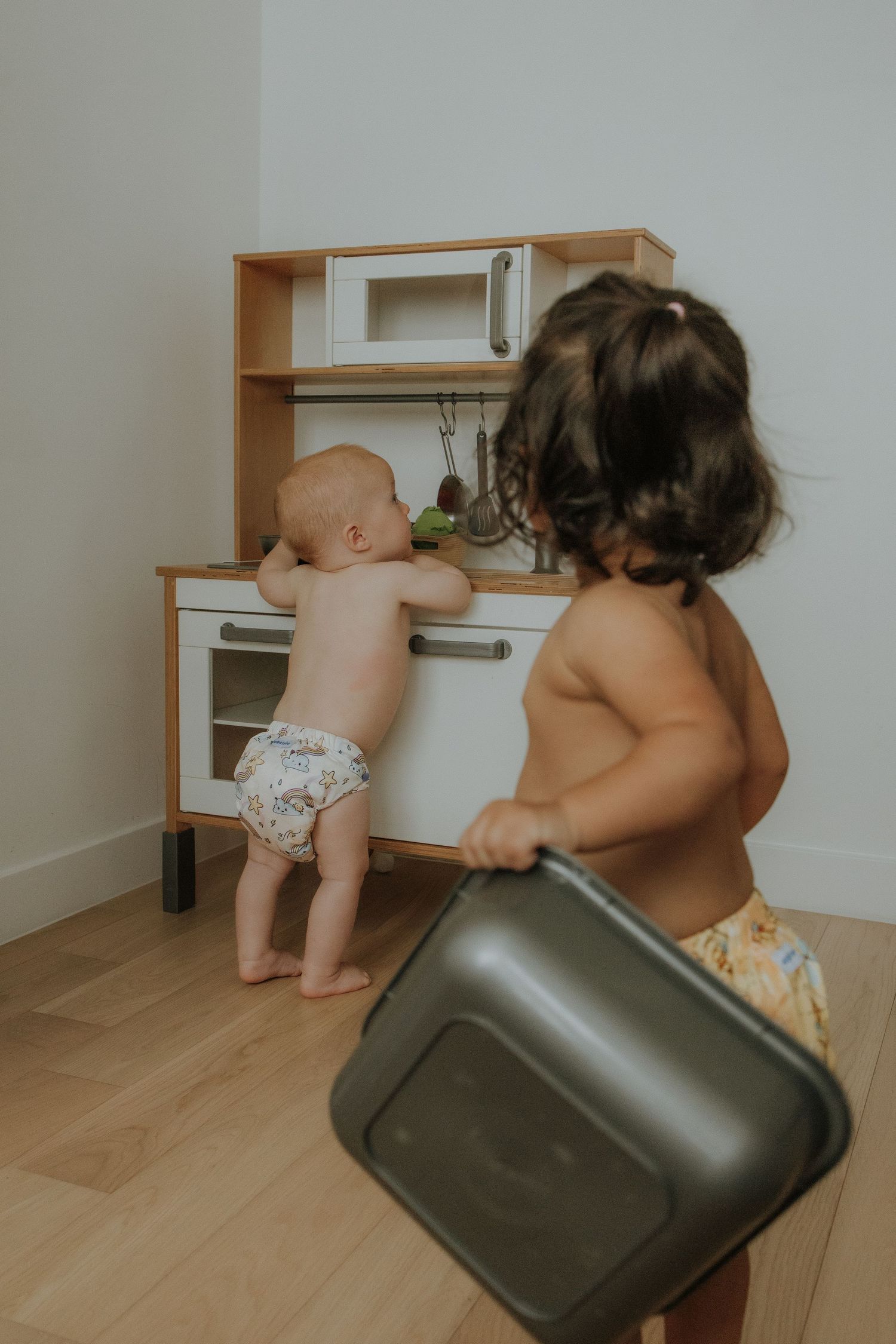 Two toddlers playing with a wooden toy kitchen, one holding a baking pan and one reaching for toy utensils.