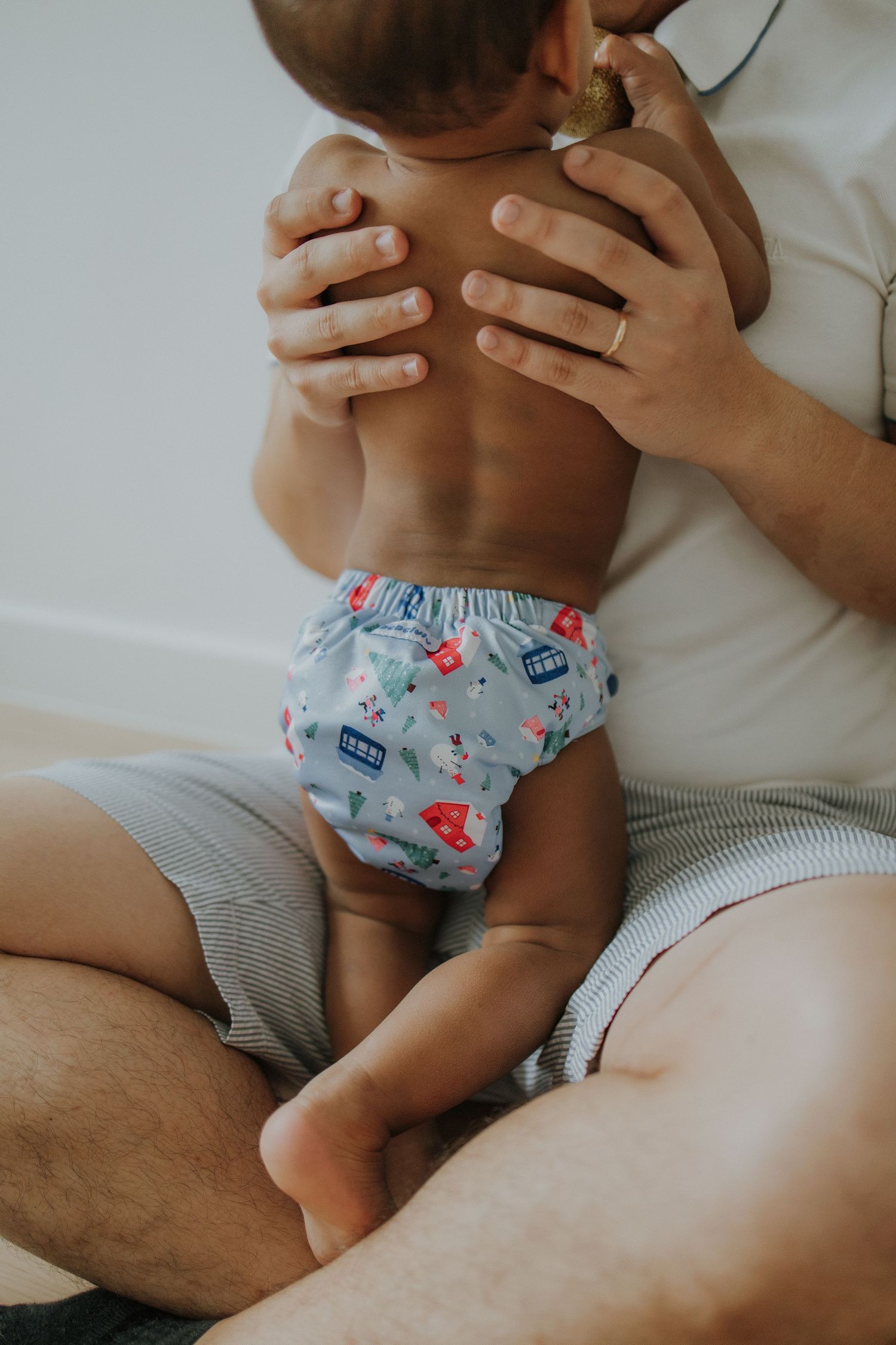 Baby wearing a colorful cloth diaper sits on parent's lap, showing bare back while being held securely.