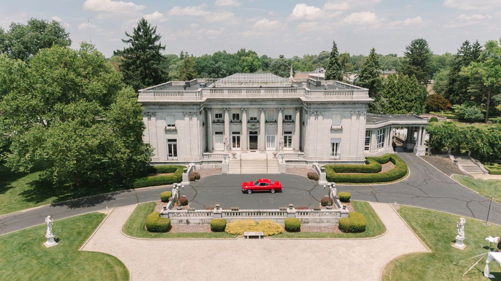 Aerial view of a grand neoclassical mansion with white columns, manicured hedges, and a red car parked in the circular driveway.