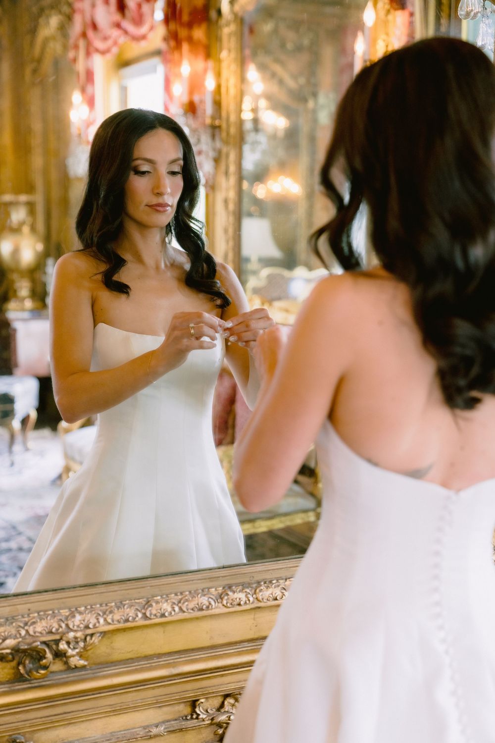 Dark-haired bride in strapless white wedding gown adjusting dress while gazing into ornate gold-framed mirror.