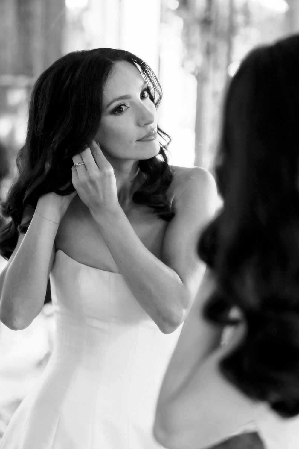 Elegant bride with dark wavy hair adjusts her earring while looking in a mirror in black and white.