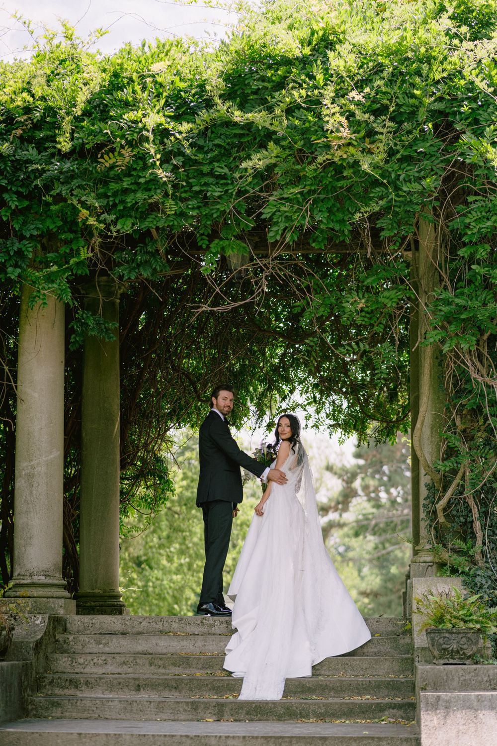 Bride and groom stand on stone steps under lush green ivy-covered pergola columns in romantic outdoor setting.