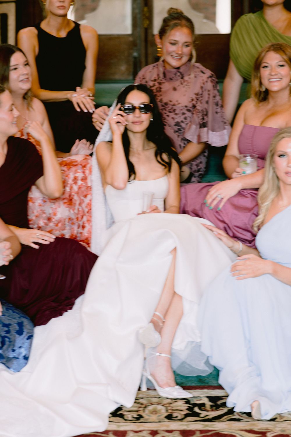 Bride in white strapless gown wearing sunglasses poses playfully with bridesmaids in colorful dresses indoors.