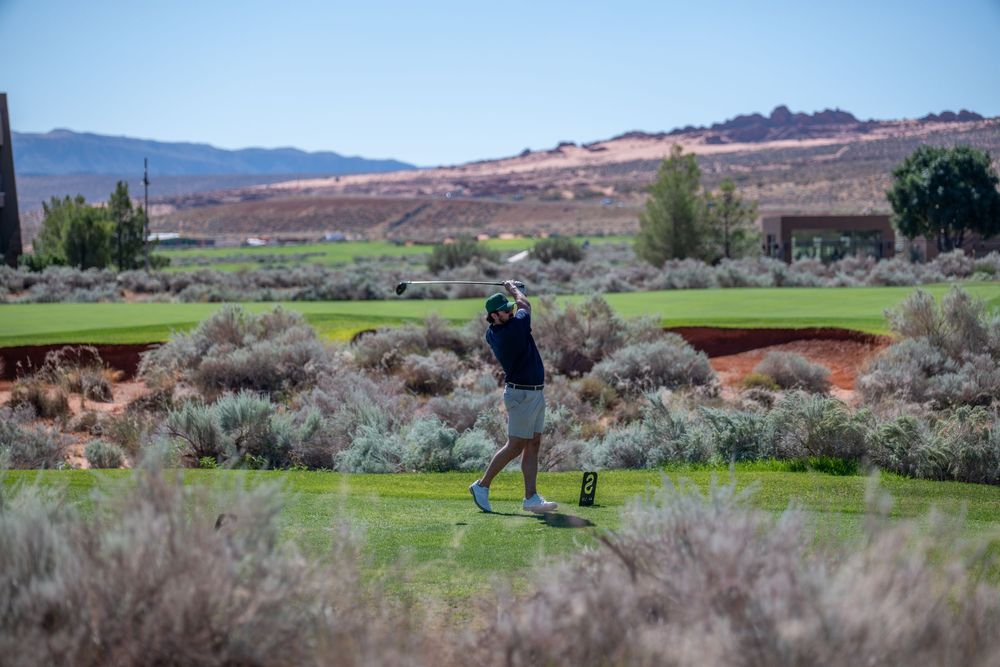 Golfer swinging a club on a scenic desert golf course with red rock formations and mountains in the background.