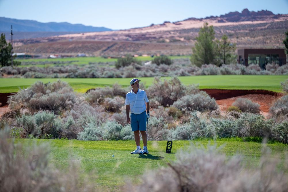Golfer standing on a desert golf course tee box surrounded by sagebrush with red rock mountains in the background.