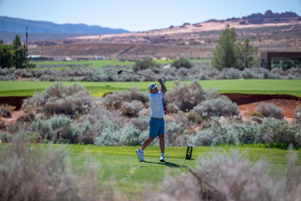 Golfer swinging club on lush green course surrounded by desert shrubs and red rock mountains in background.