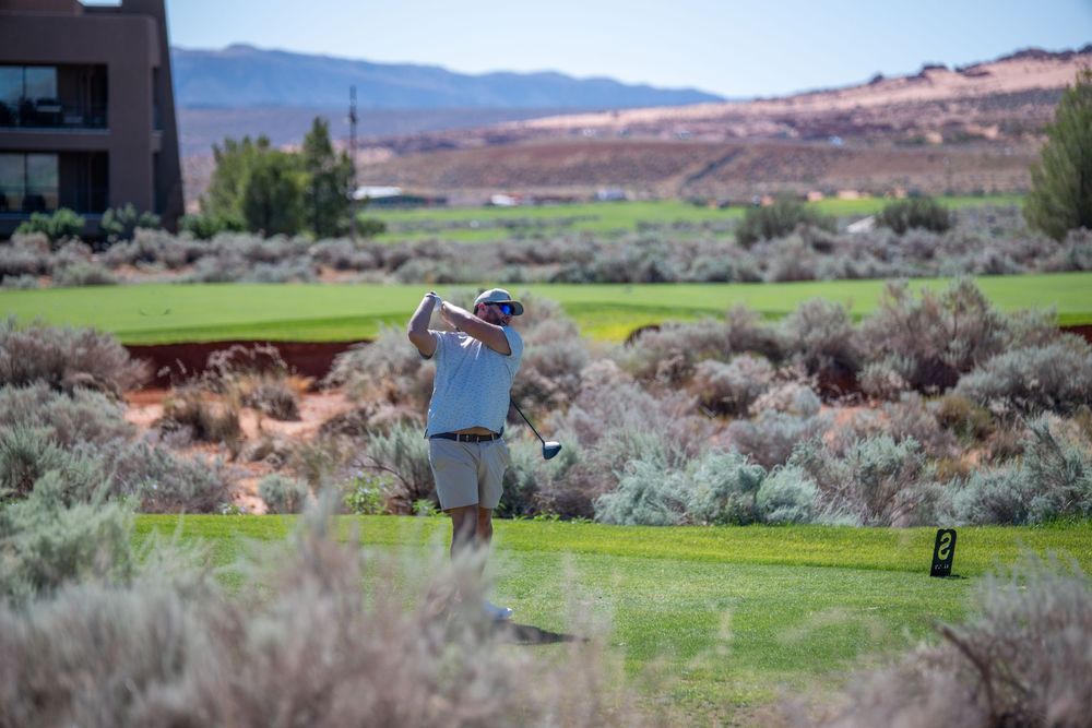 Golfer mid-swing on a scenic desert golf course with sagebrush, red rock mountains, and a building in the background.