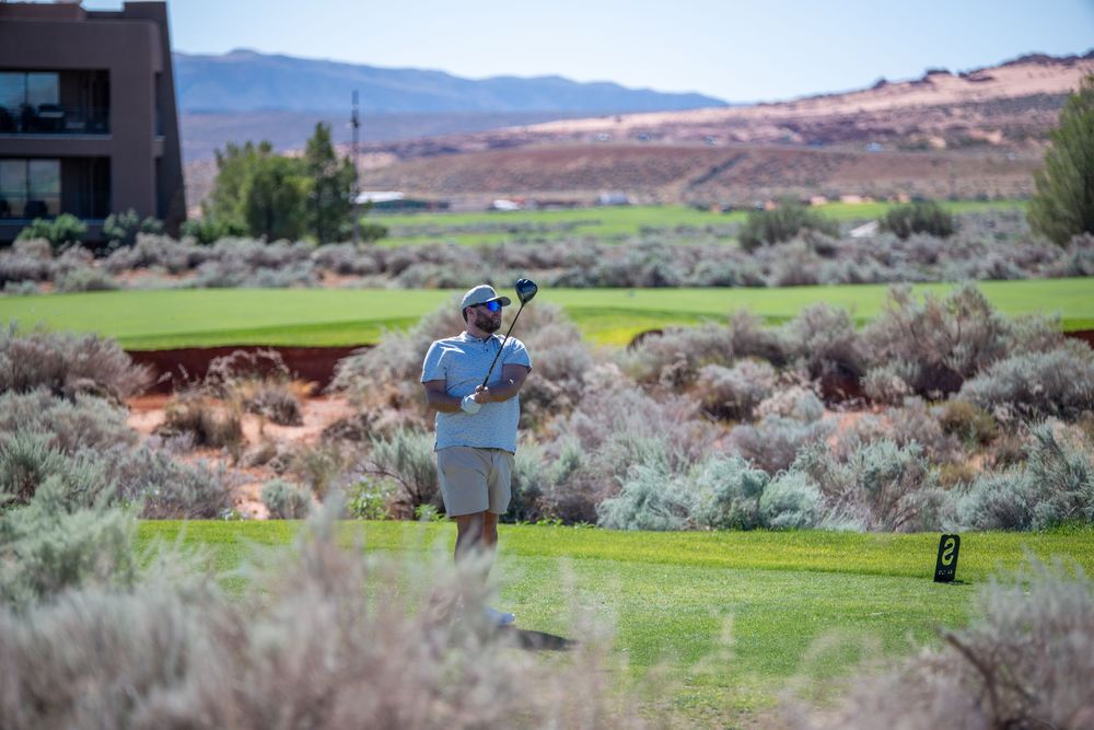 Golfer swinging club on scenic desert golf course with red rock mountains and sagebrush in background.
