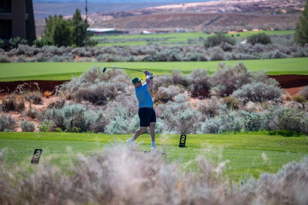 Golfer mid-swing on a lush green course surrounded by desert shrubs and red rock landscape in St. George, Utah.