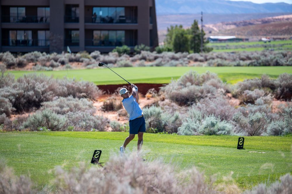 Golfer swinging a club on a lush green course with desert shrubs and a modern building in the background.