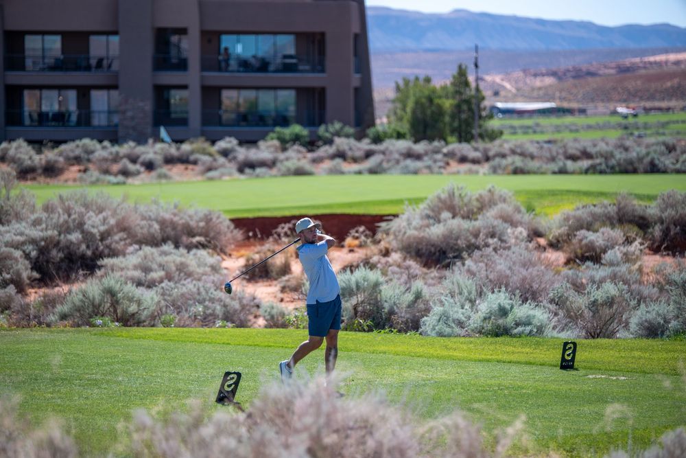 Golfer swinging on a lush green course with desert shrubs and a modern building backdrop in a scenic arid landscape.