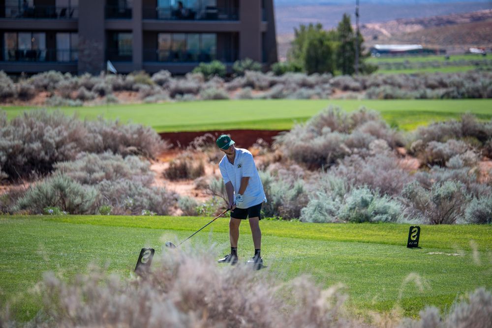 Golfer teeing off on a lush green golf course surrounded by desert sagebrush with a modern building in the background.