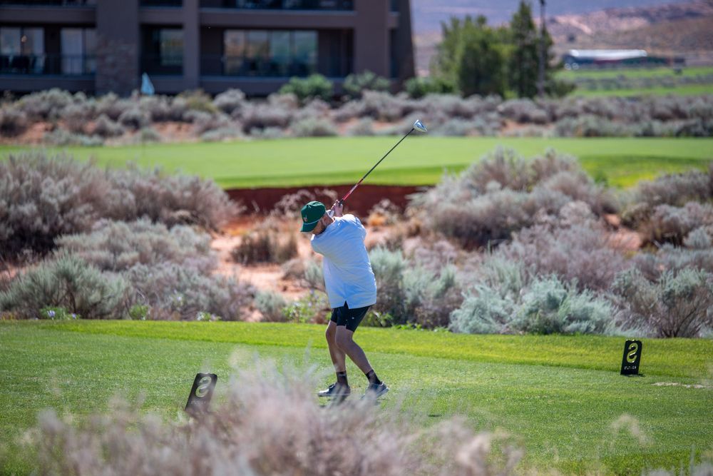 Golfer completing a swing on a lush golf course surrounded by desert shrubs and a modern building backdrop.