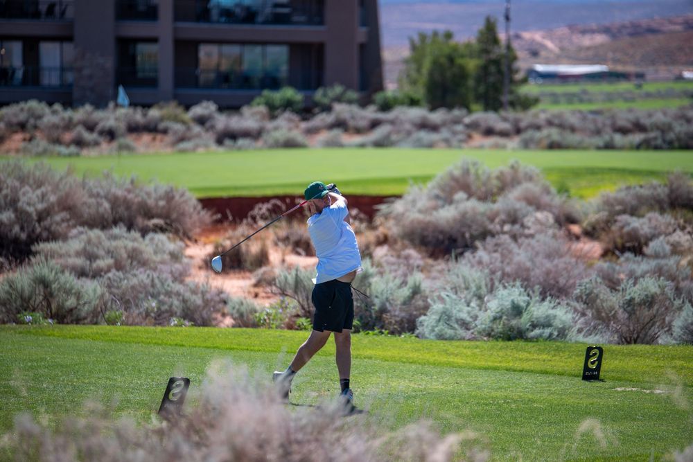 Golfer swinging club on lush green course with desert shrubs and modern building in background.