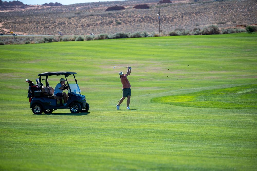 Golfer completing a swing on a sunny green fairway beside a blue golf cart with arid hills in the distance.