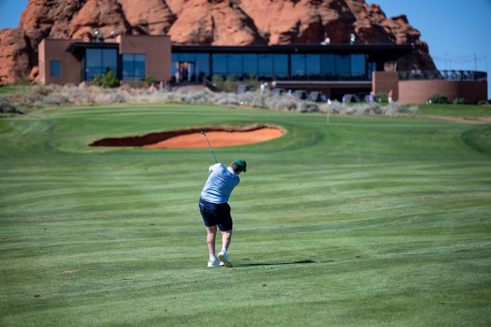 Golfer swinging a club on a lush green course with red rock formations and a clubhouse in the background.