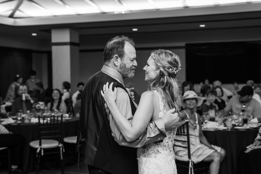 Black and white photo of bride and groom sharing first dance at wedding reception with guests seated in background.