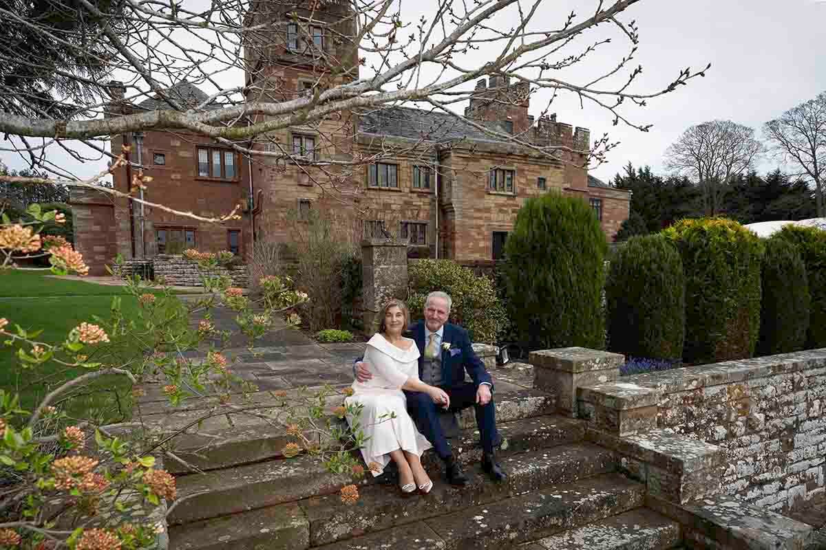 Couple sitting on stone steps outside a grand historic red brick manor house surrounded by lush gardens.