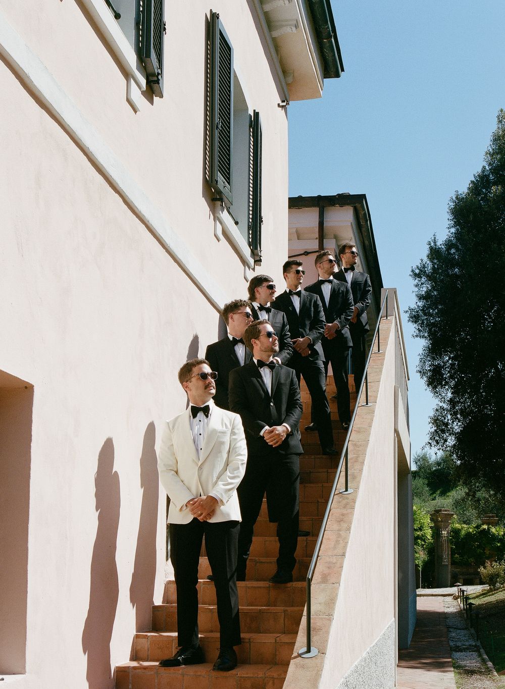 Groom in white tuxedo jacket stands with groomsmen in black tuxedos on outdoor stone steps at a sunny wedding venue.