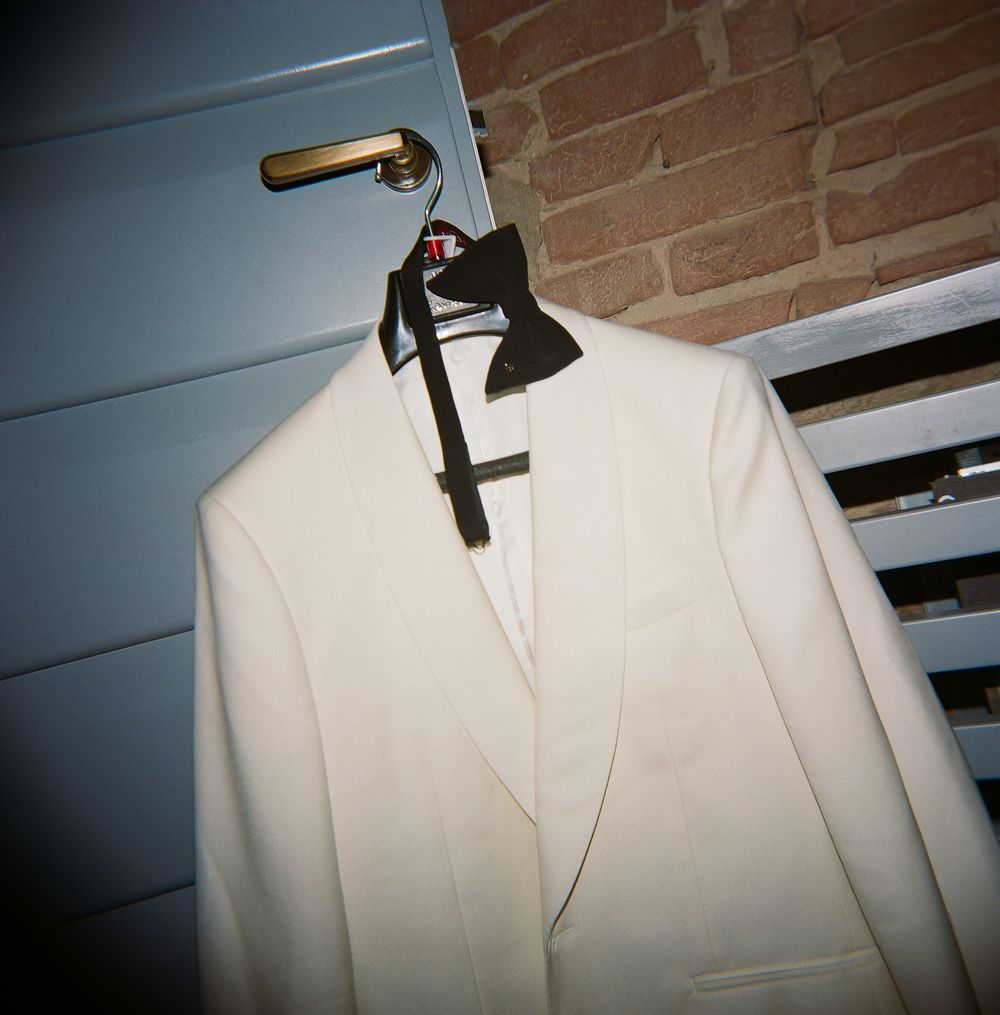 White tuxedo jacket with black bow tie displayed on a hanger against a brick wall and blue door.