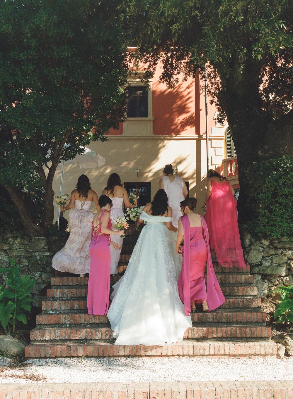 Bride in white gown walks up brick steps with bridesmaids in pink dresses toward a sunlit villa entrance.