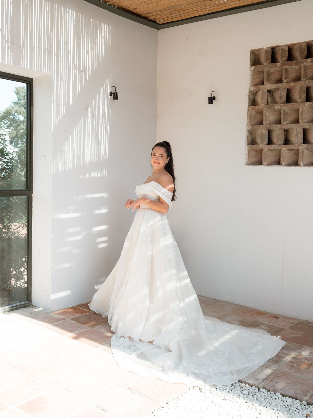 Bride in elegant off-shoulder white wedding gown with long train poses in sunlit white venue.