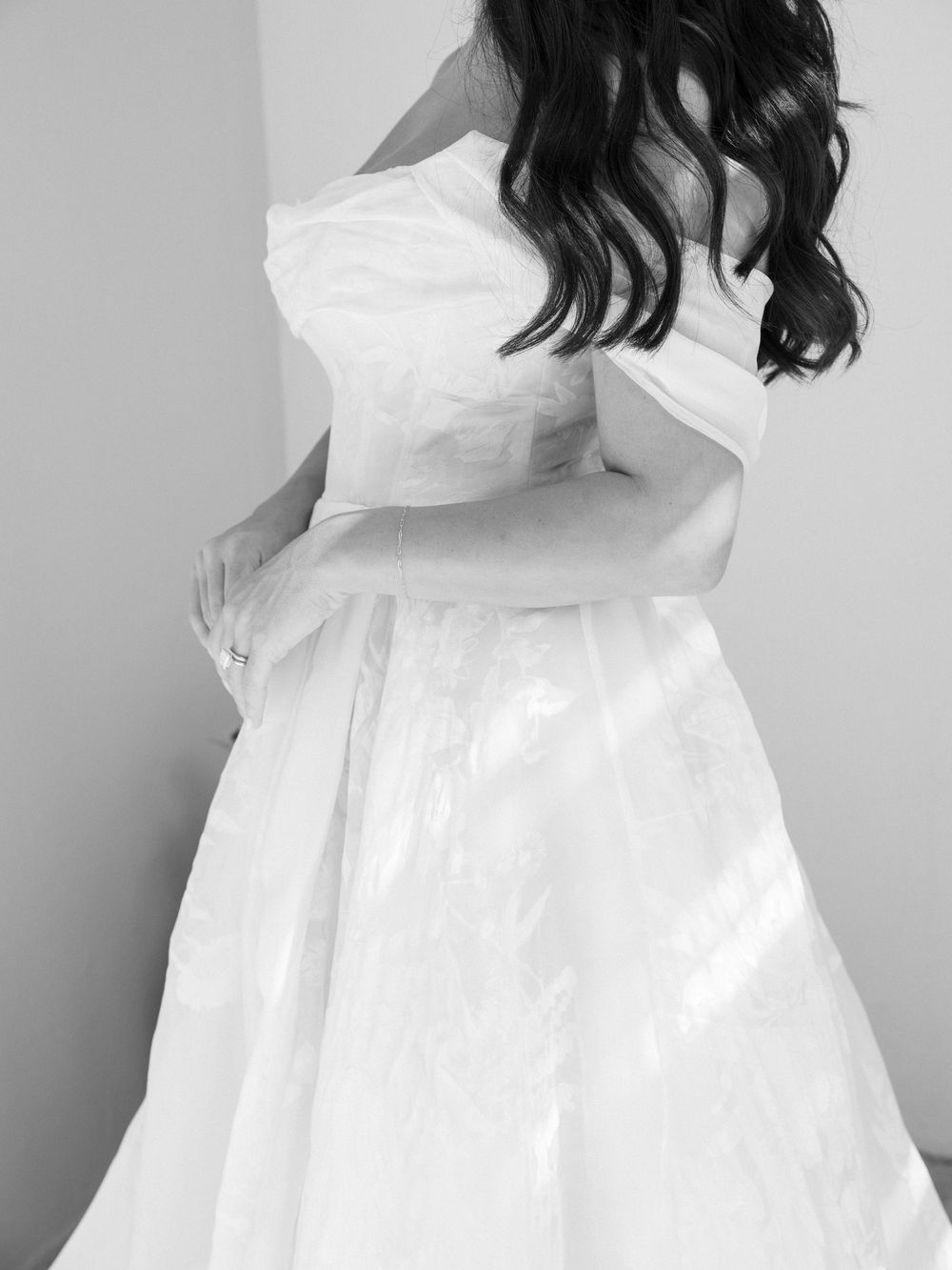 Black and white photo of bride in off-shoulder wedding gown adjusting her dress.