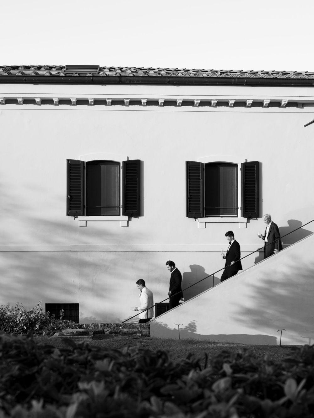 Black and white photo of four men in suits descending outdoor stairs beside a classic European building with shuttered windows.