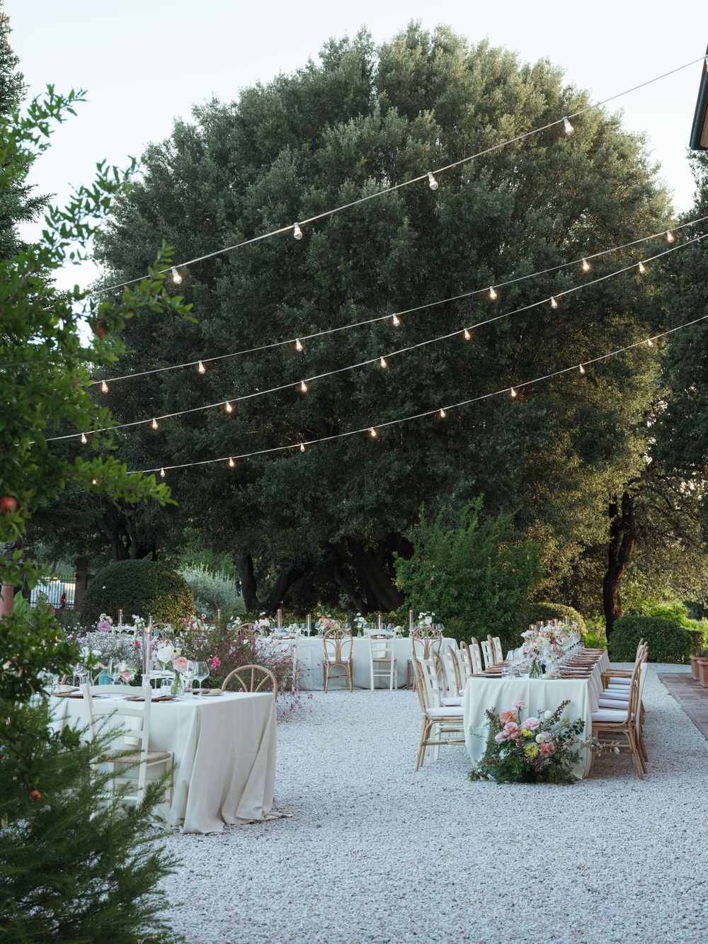 Elegant outdoor wedding reception with string lights, white tablecloths, and floral centerpieces on a gravel garden patio.