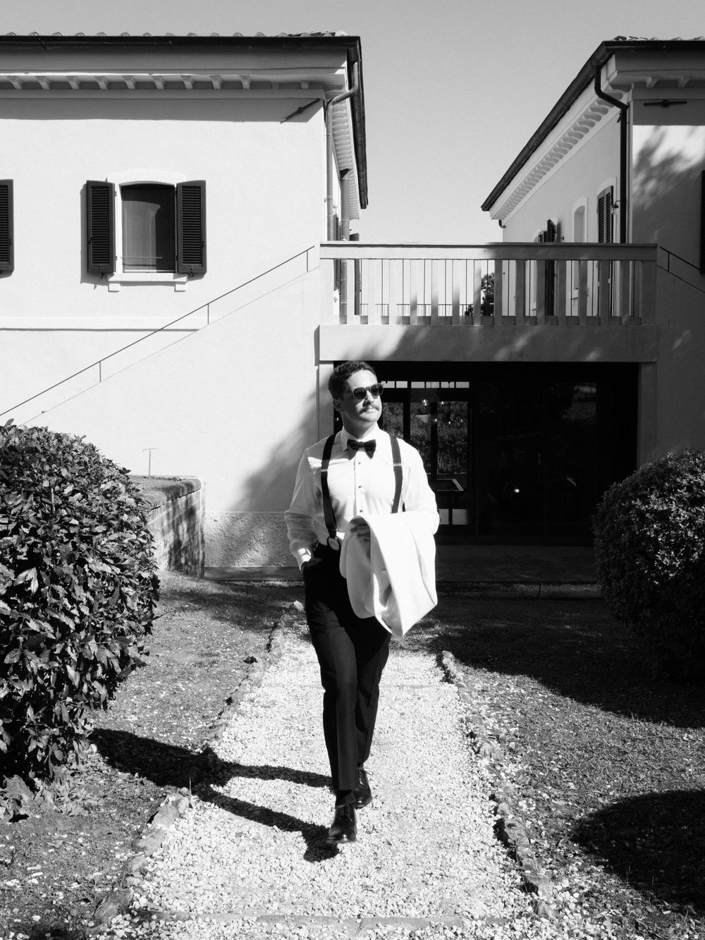 Stylish person in black and white outfit walks gravel path near Italian villa in B&W photo.