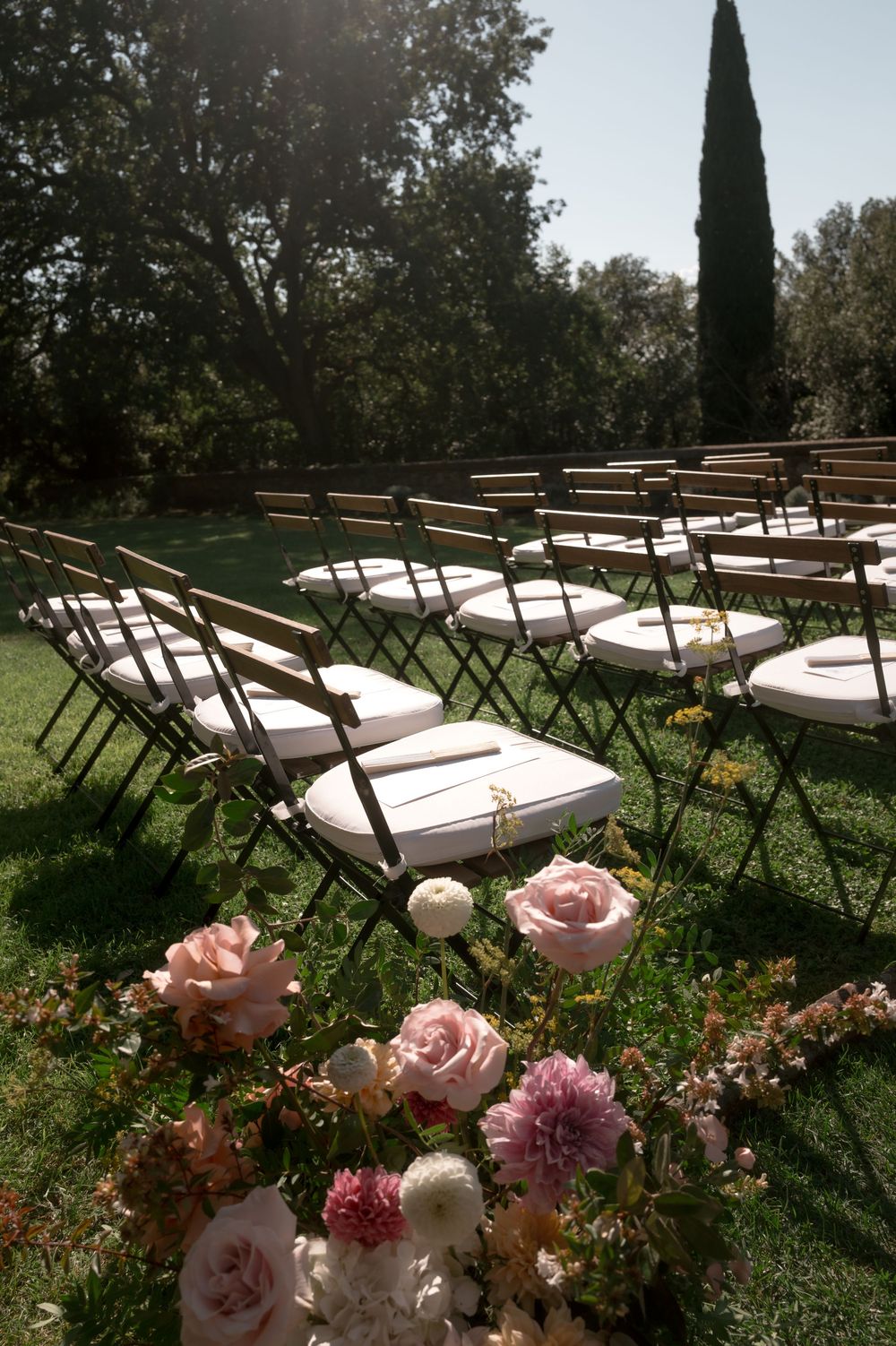 Outdoor wedding ceremony setup with white cushioned folding chairs on green lawn, pink floral arrangements in foreground, cypress tree backd