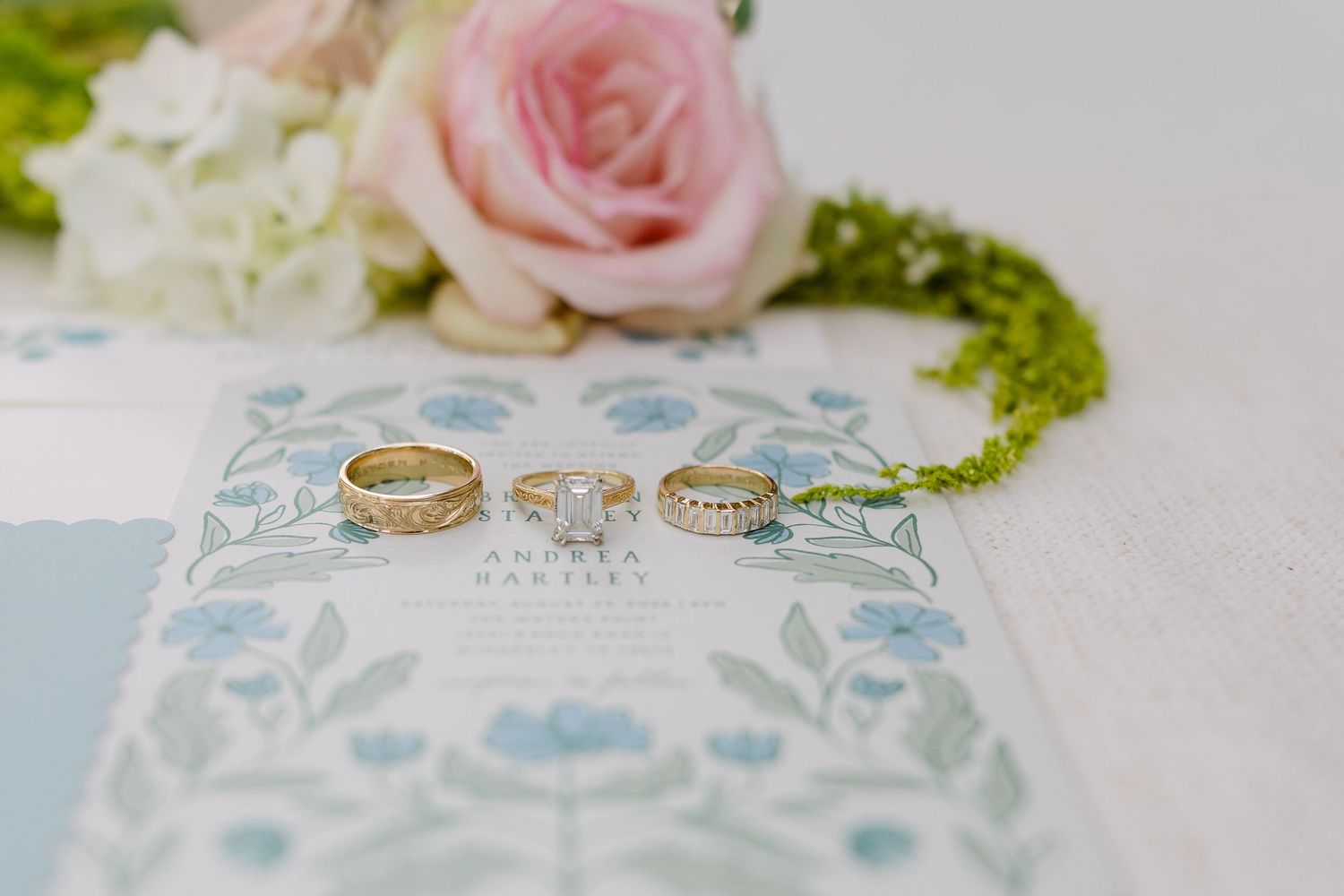 Gold wedding bands and diamond engagement ring displayed on floral wedding invitation with pink roses in background.