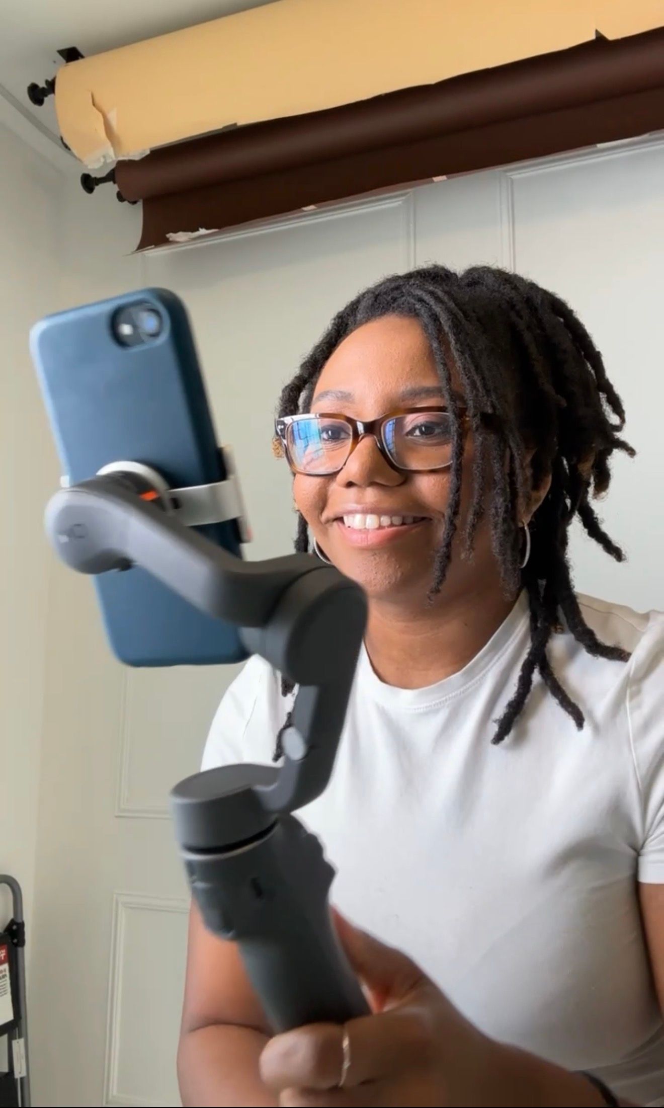 Smiling woman with locs and glasses holding a smartphone gimbal stabilizer in a white room.