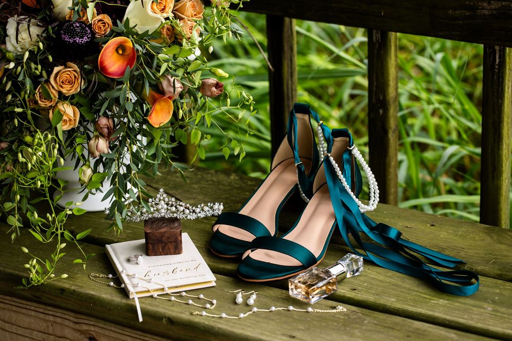 Teal wedding heels with pearl ankle straps beside a floral bouquet, perfume, and jewelry on a wooden surface.