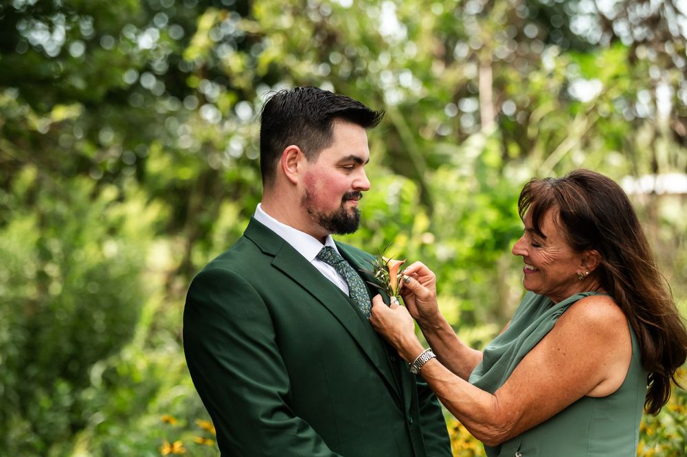 A woman in a green dress pins a boutonniere on a groom in a dark green suit outdoors.