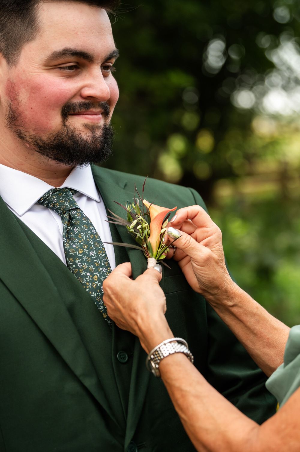 Groom in dark green suit and floral tie having boutonniere pinned to lapel outdoors at wedding.