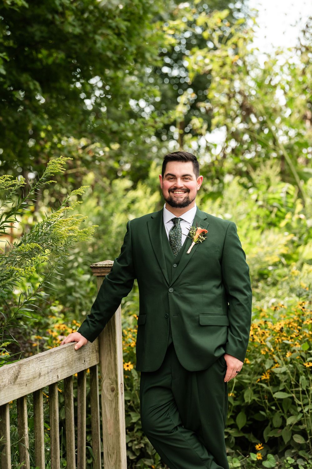 Smiling groom in dark green three-piece suit with floral boutonniere posing by wooden fence in lush garden setting.